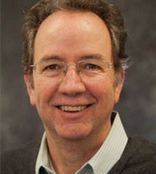 Smiling man with glasses, wearing a gray sweater over a collared shirt, against a blurred background.
