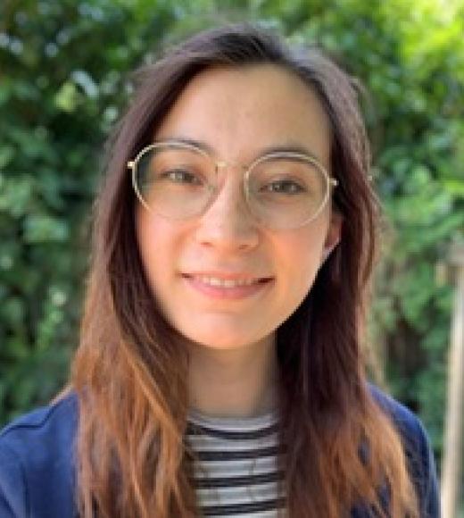 A young woman with glasses smiles in front of green foliage.